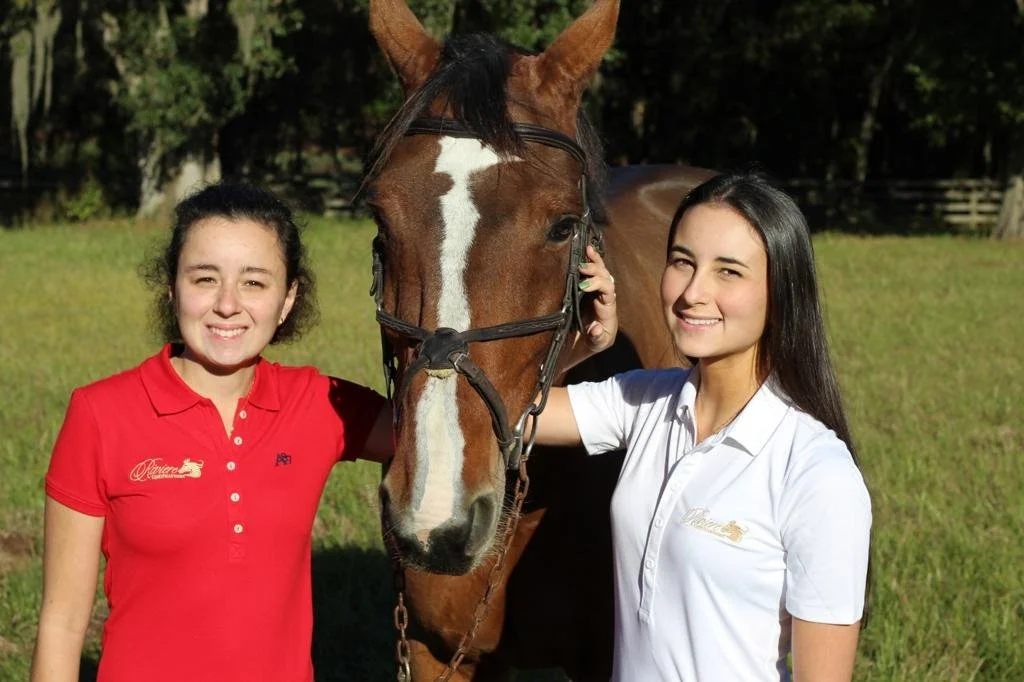 Two women in polo shirts smiling next to a brown horse with a white stripe on its face, in a grassy field with trees in the background.