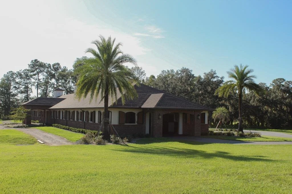 A single-story house with a dark roof, brick and white exterior, surrounded by palm trees and a grassy lawn under a blue sky.