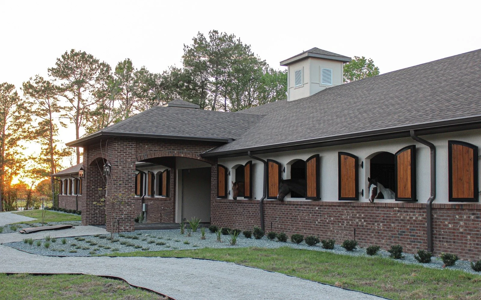 A barn with sliding wooden shutters on the windows, horse heads seen peeking out, brick and white exterior, located outdoors at sunset with trees in the background.