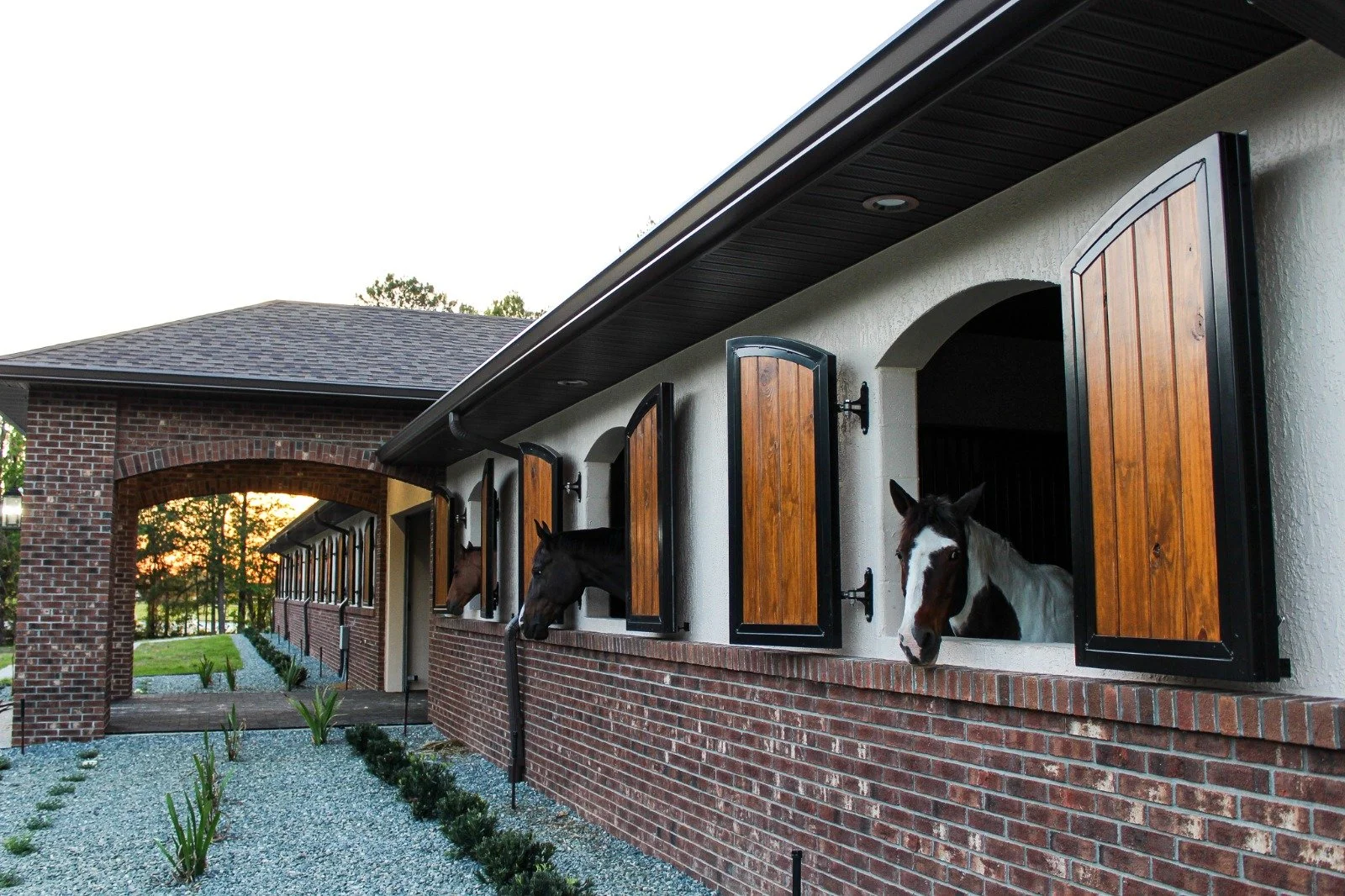 Horses looking out of windows in a brick stable building at sunset.