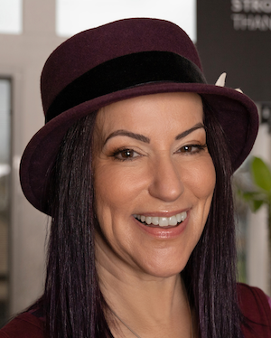 A woman with glasses and long, gray and brown hair smiling at the camera, sitting at a table in an indoor setting.