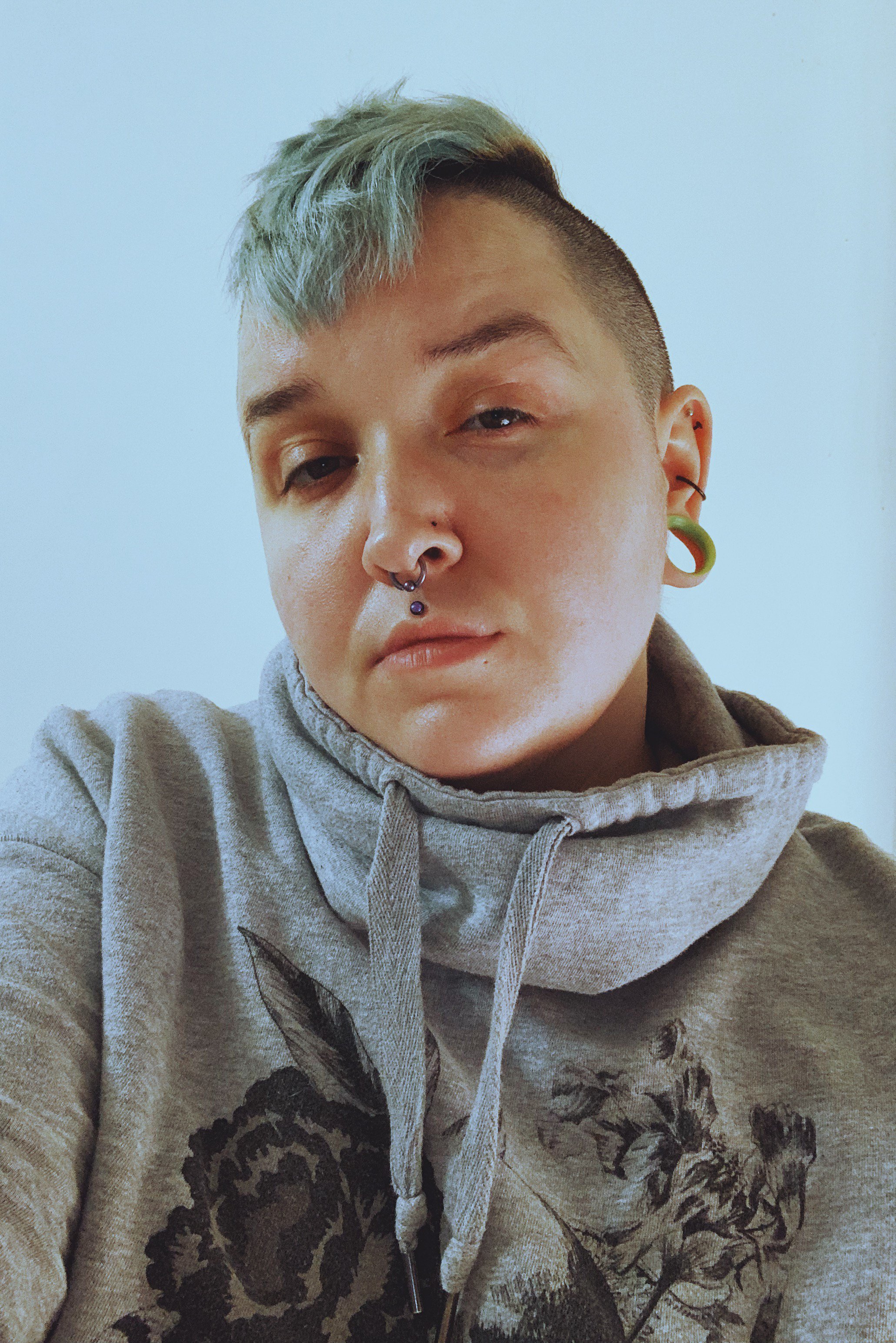 A person with short green hair, multiple facial piercings, and wearing a gray hoodie, taking a selfie in front of a plain white wall.