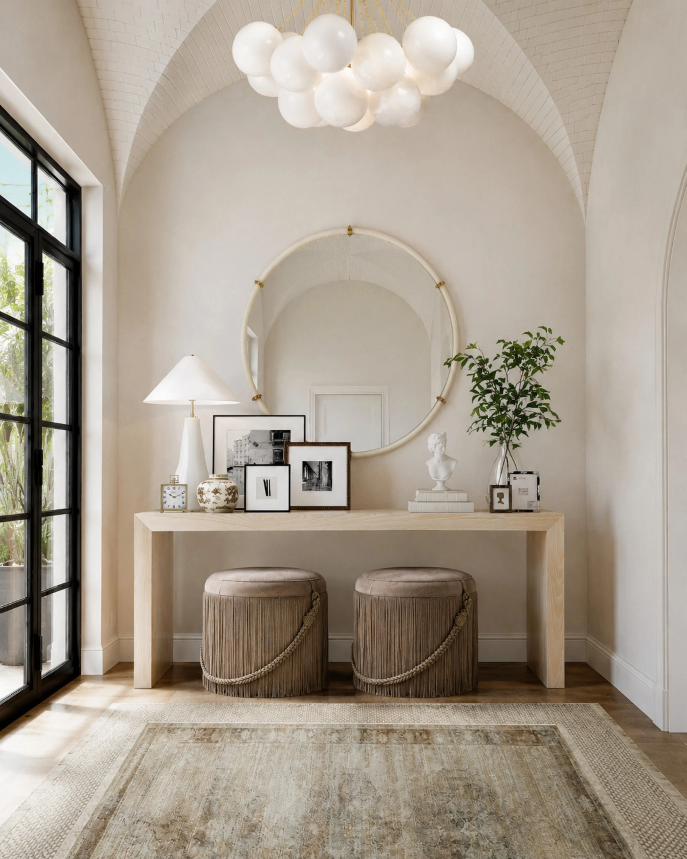 Classic entryway, but make it feel collected. I love the mix of the pale wood console, oversized round mirror, sculptural lighting, layered artwork, and fringe ottomans tucked underneath. It feels elegant without being too formal, and the soft neutra