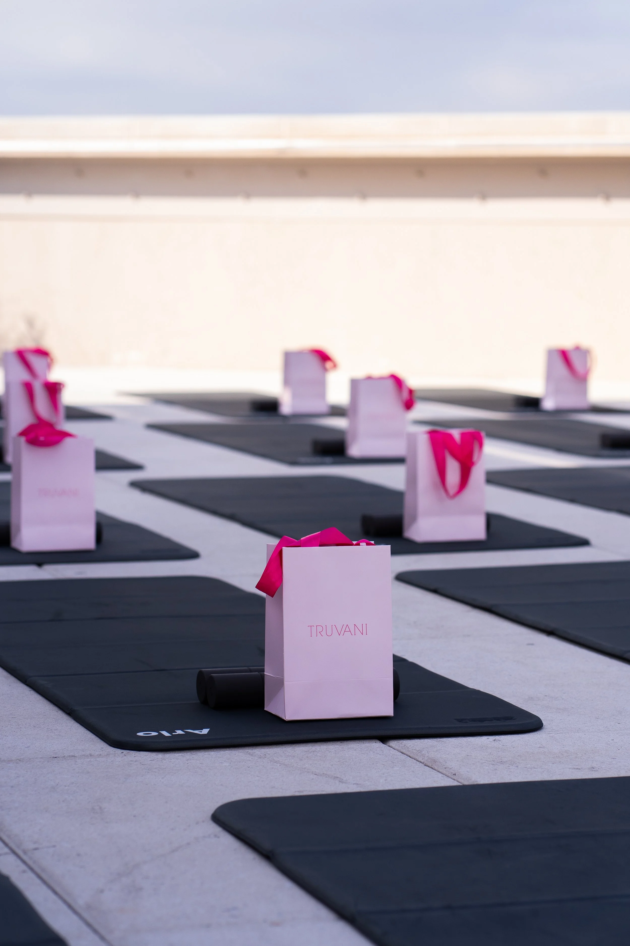 Yoga mats on a rooftop with pink gift bags labeled 'TRUVANI' placed on each mat, arranged in a grid pattern.