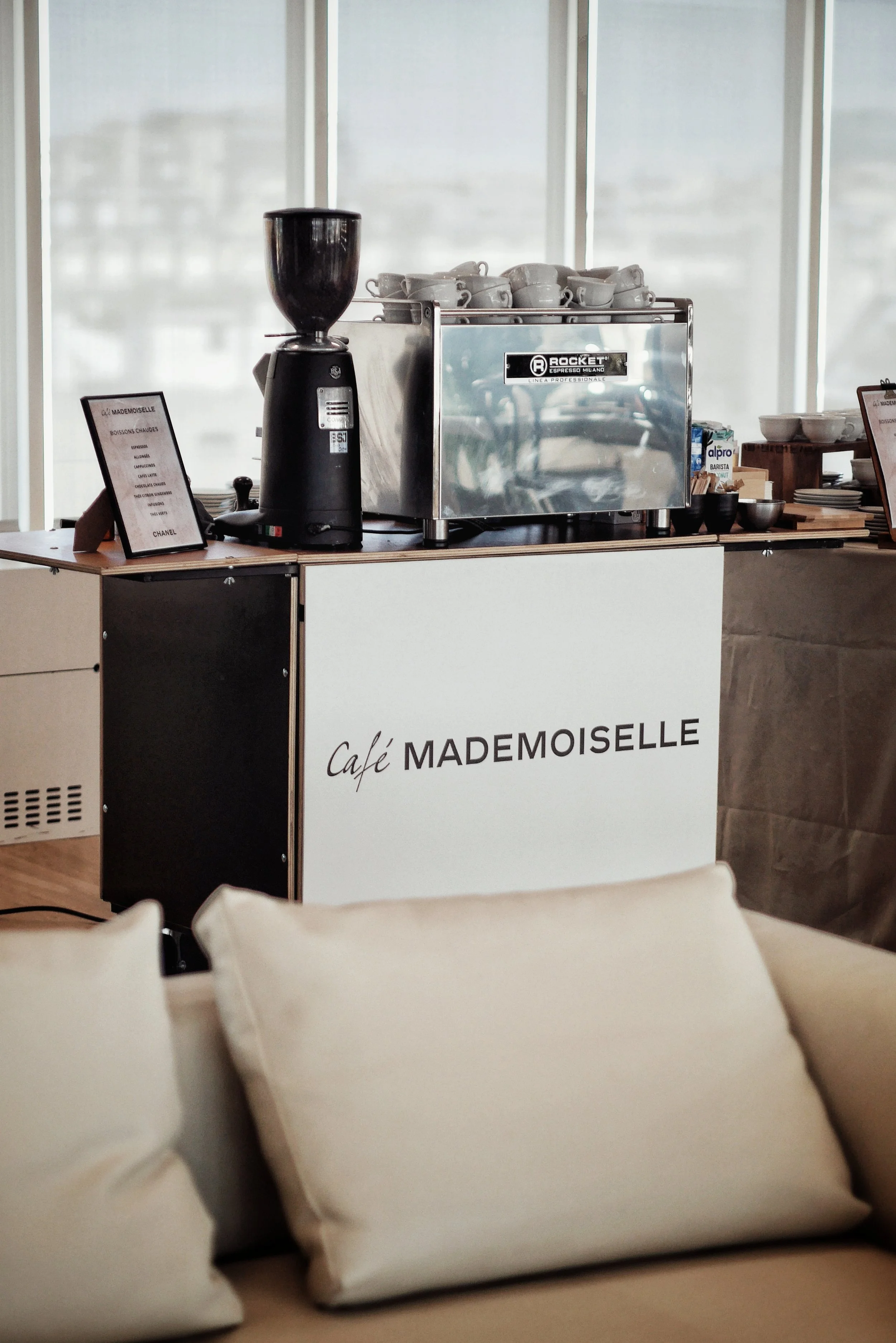 Une station de café avec une machine à espresso, un moulin à café, des tasses et des ustensiles, dans un cadre intérieur moderne. Le panneau indique 'Café Mademoiselle'.