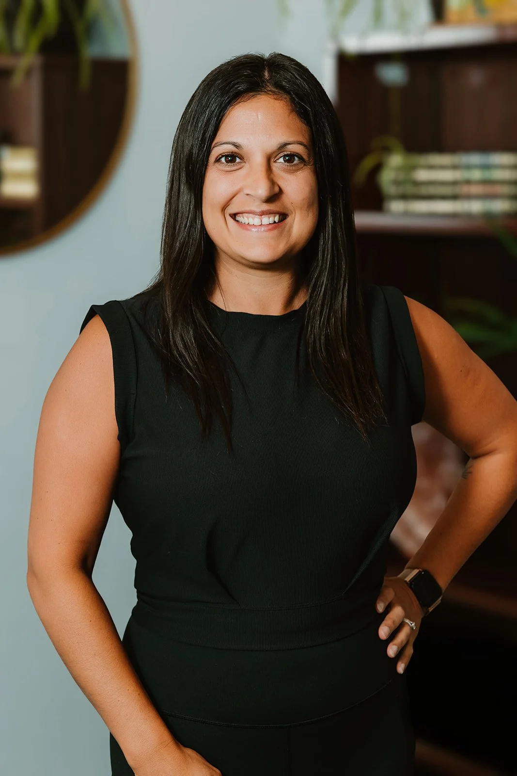 A woman with long dark hair smiling, wearing a black sleeveless top, standing indoors near a bookshelf and a round mirror.