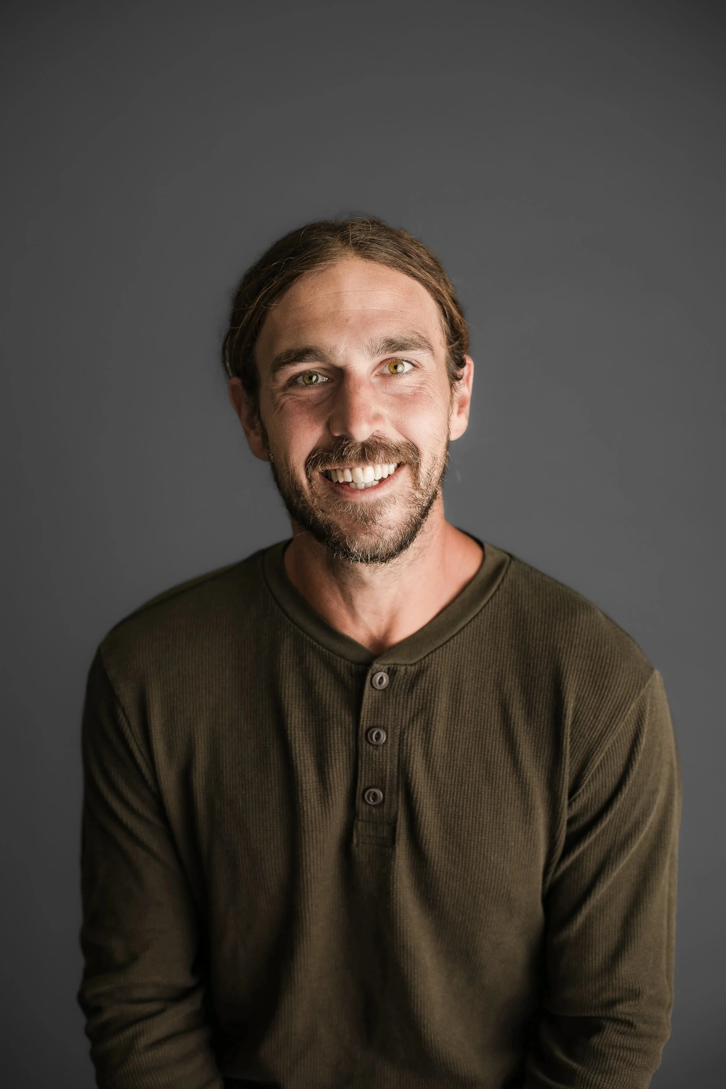 A smiling man with a beard and long hair, wearing a green long-sleeve shirt, against a gray background.