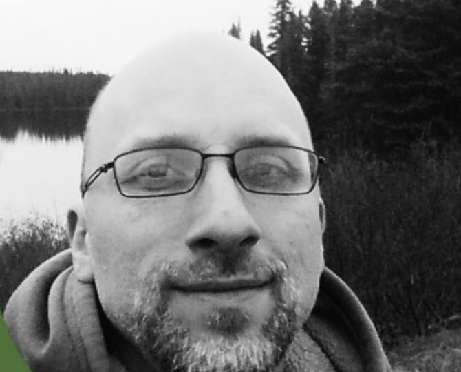 Un homme avec lunettes et barbe dans un environnement naturel avec de l'eau et des arbres en arrière-plan.