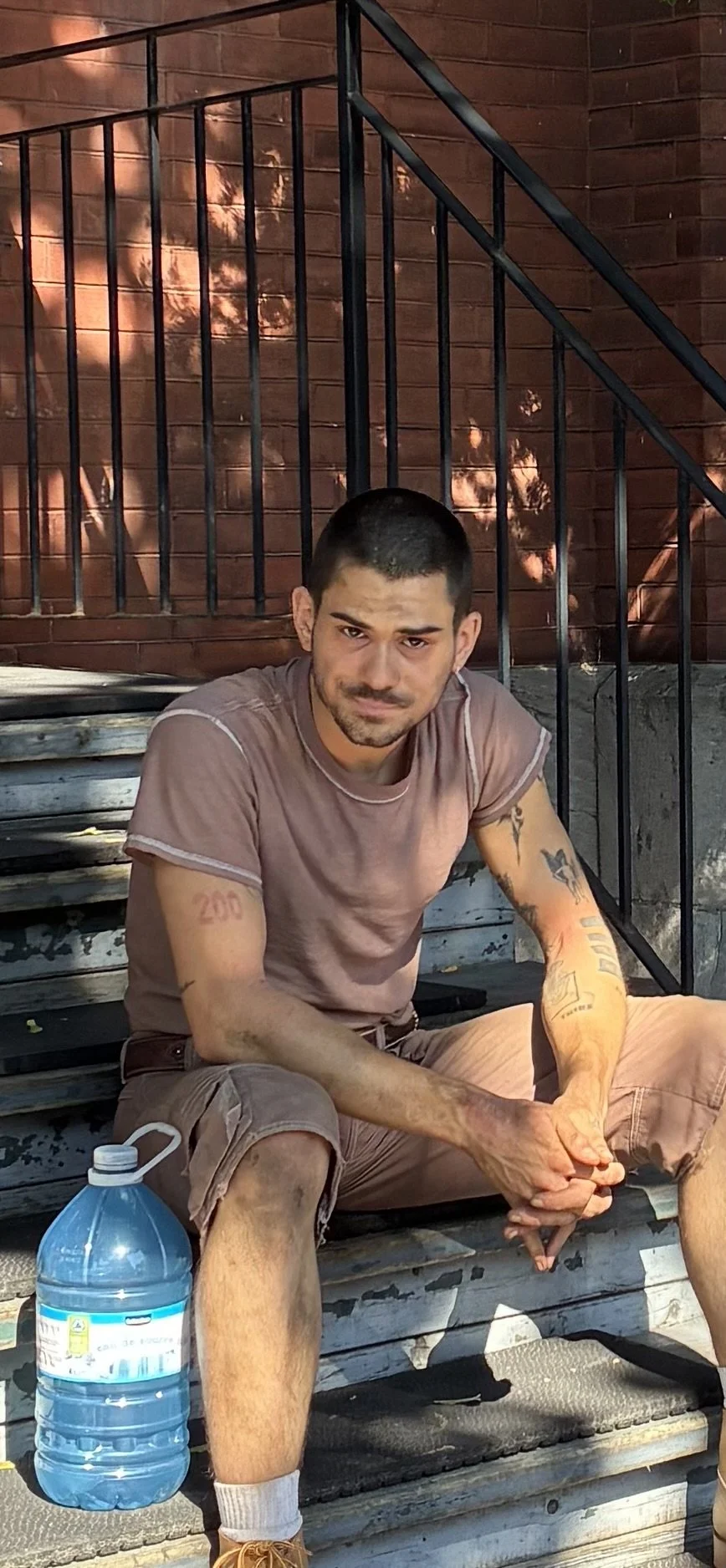 Un homme assis sur un banc en bois, portant un t-shirt marron et des shorts, avec un flacon d'eau à côté de lui, dans un environnement urbain avec un mur en briques en arrière-plan.