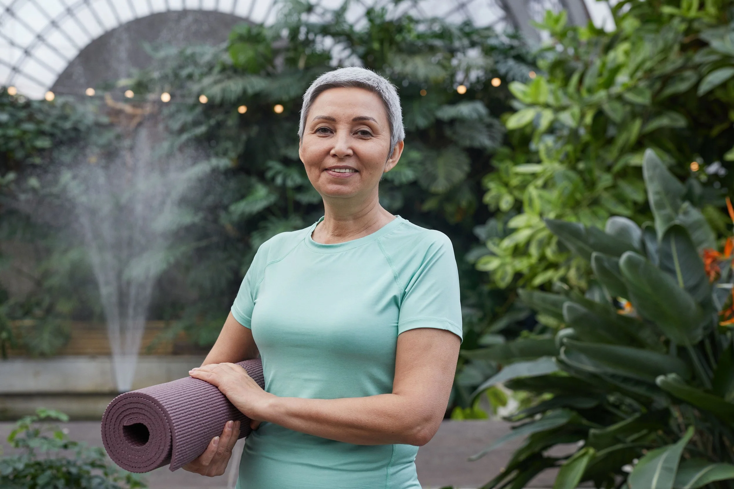 A woman with short gray hair smiling and holding a purple yoga mat in a lush greenhouse filled with green plants.