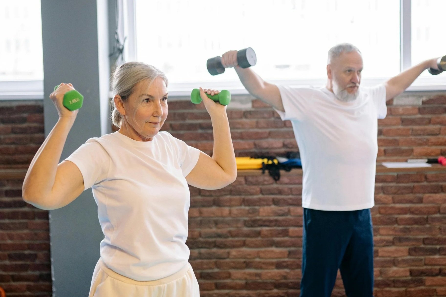 An elderly woman and man exercising indoors. The woman is lifting green dumbbells with both arms bent at the elbows, and the man is extending his arms holding black dumbbells. They are in a fitness room with a brick wall and windows in the background.