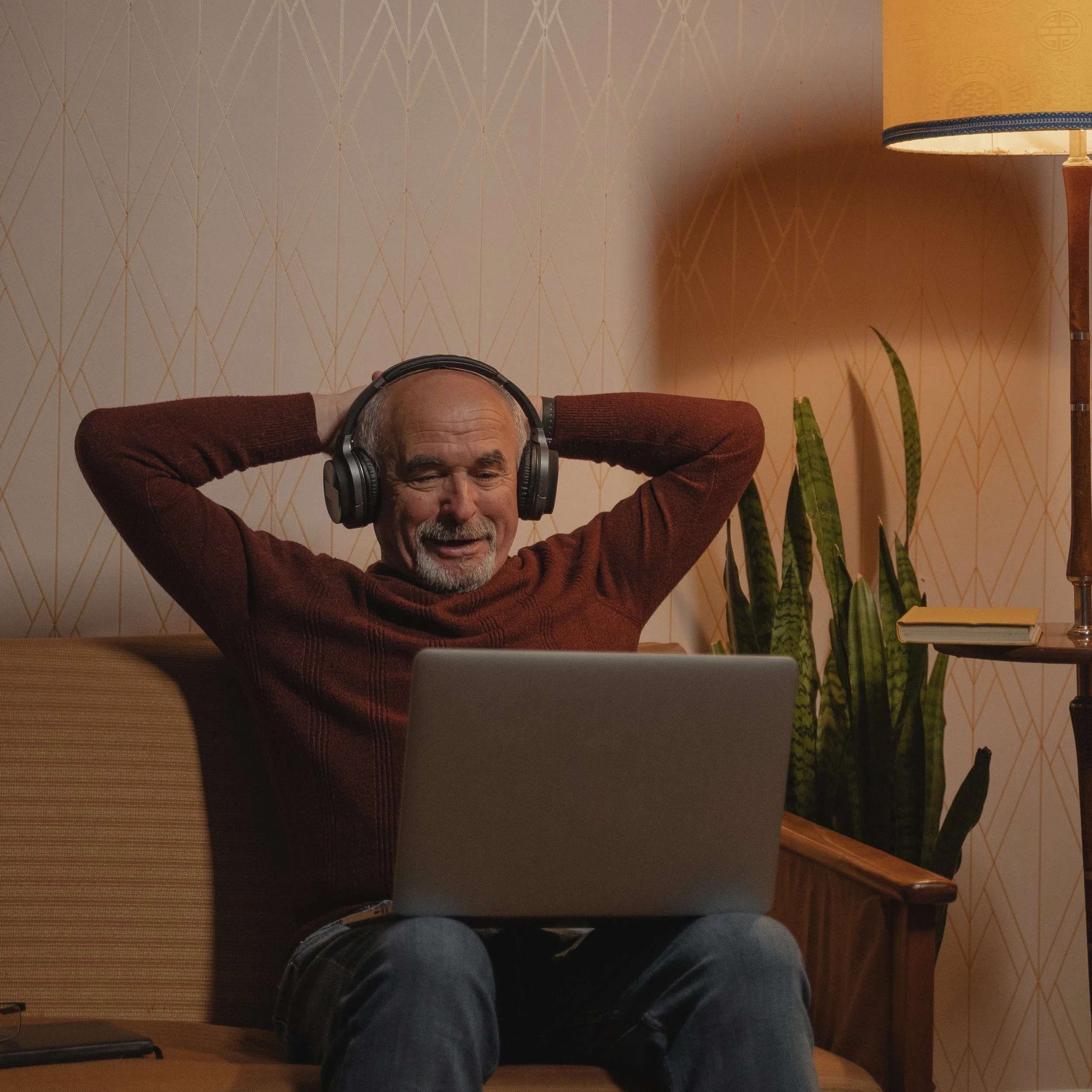 An elderly man sitting on a couch with a laptop on his lap, wearing headphones and smiling with arms behind his head in a cozy living room.