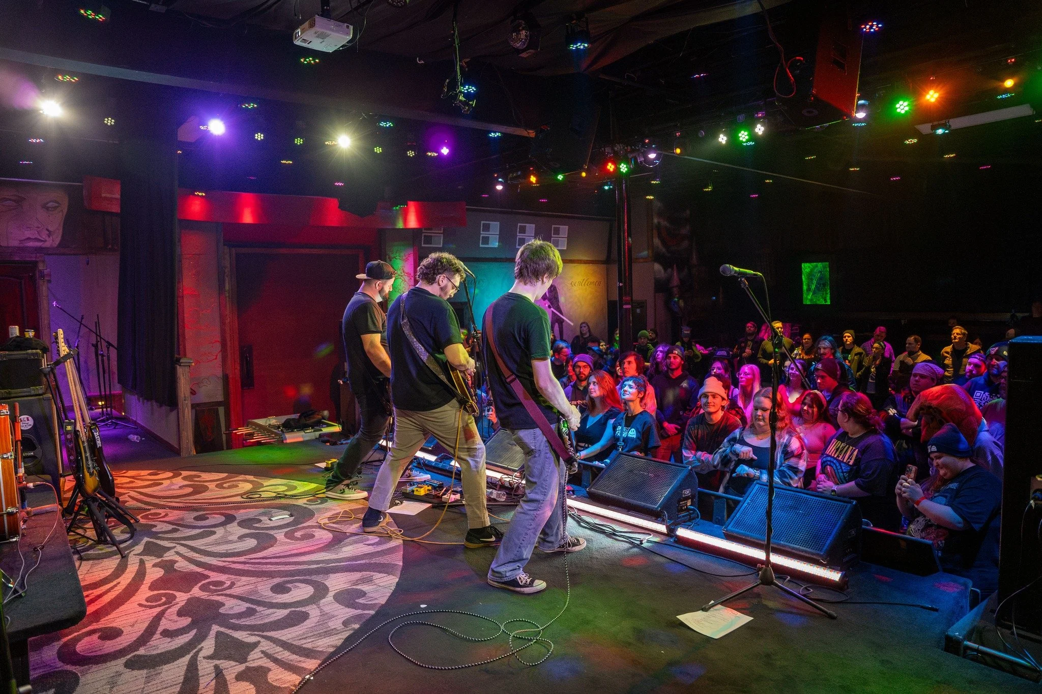 A band performing live on stage in front of an audience at a concert venue with colorful stage lighting and people enjoying the music.