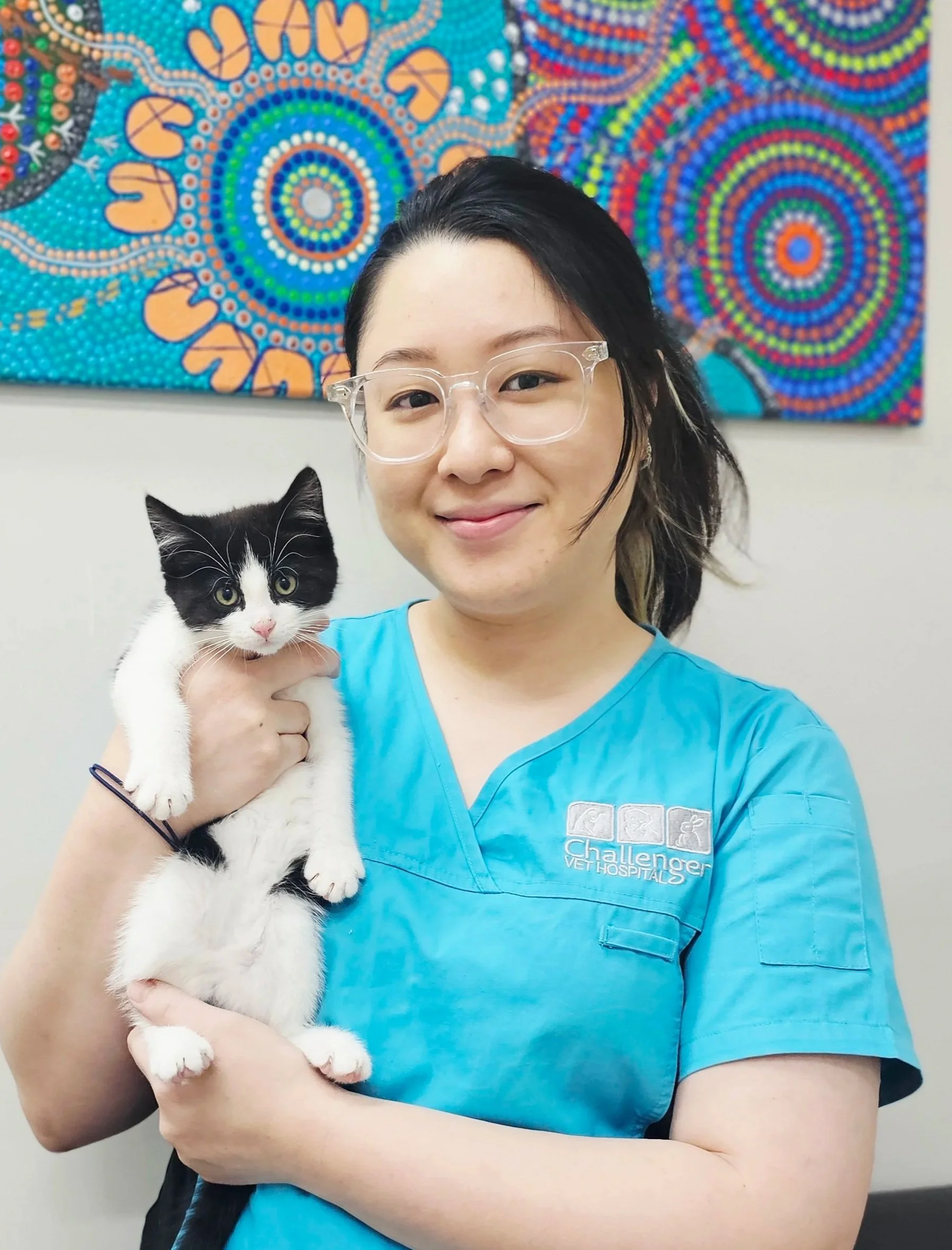 A woman wearing blue veterinary scrubs and clear glasses is holding a black and white kitten. They are standing in front of a colorful, dot-patterned artwork on a white wall.