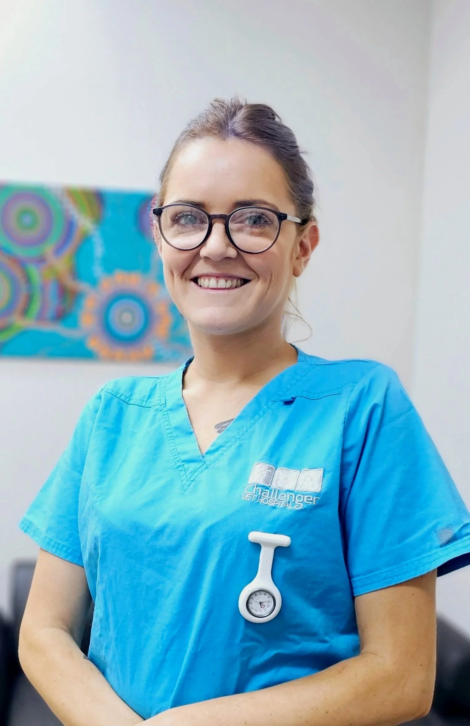 A smiling female veterinarian or medical professional wearing blue scrubs and glasses, standing in a clinic or hospital with a colorful abstract painting on the wall behind her.