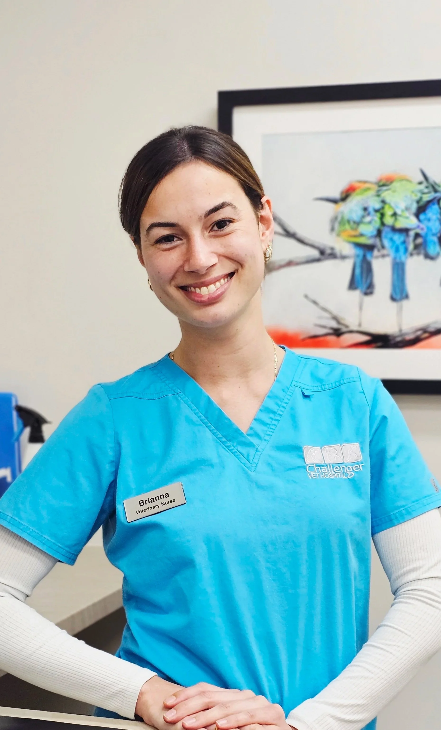 A smiling female veterinary nurse in blue scrubs standing in front of a framed colorful bird painting.