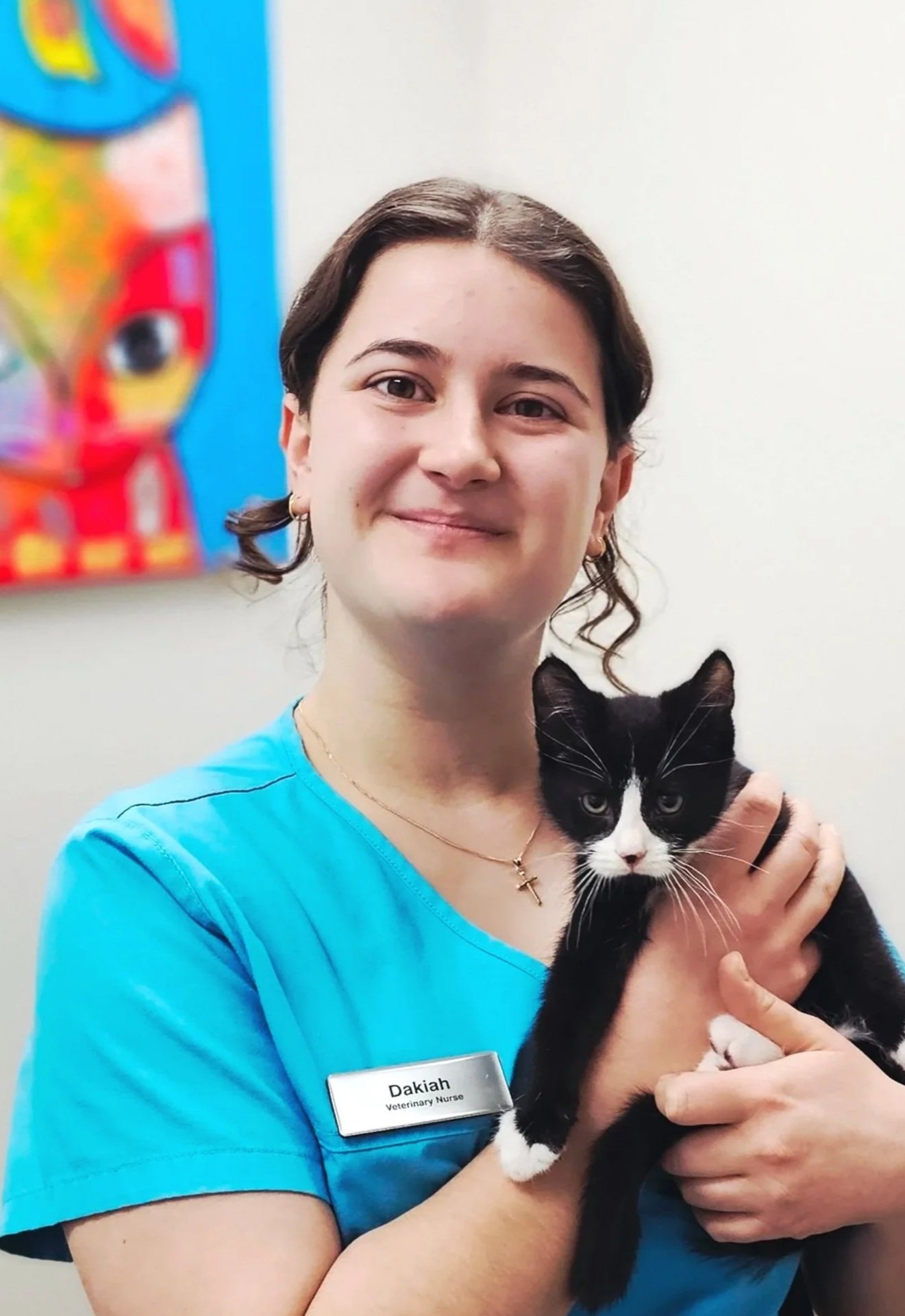 A smiling woman in a blue veterinary nurse uniform holding a black and white cat in her arm, with an abstract colorful painting on the wall in the background.