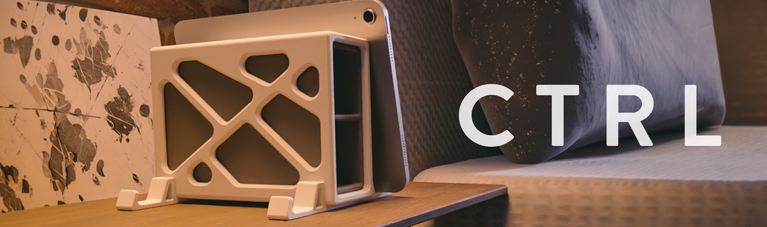 A close-up of a desk with a tablet, a decorative white geometric stand, a laptop with a black and gray textured cover, a piece of printed map with trees and leaves, a black-and-white patterned cushion, and a white bed or mattress in the background. The word "CTRL" is overlaid on the image.