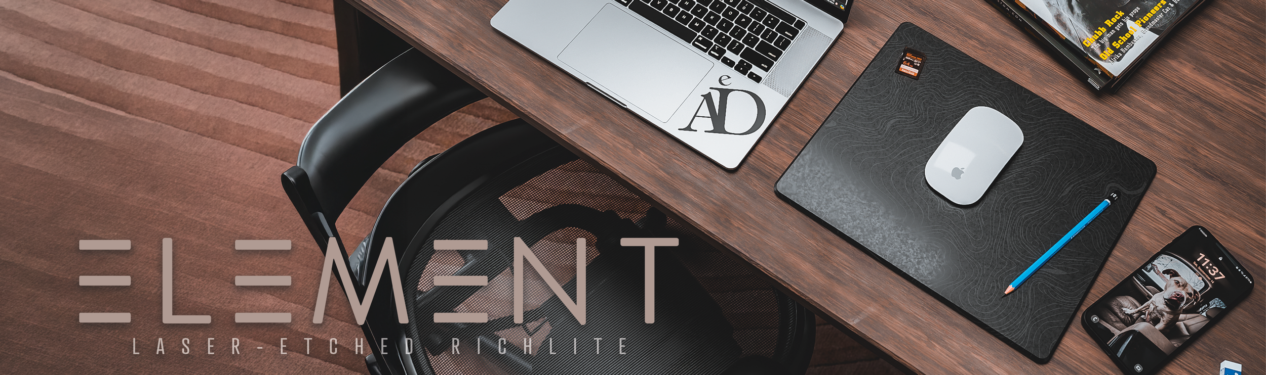 An organized desk with a laptop, a mouse on a black textured mouse pad, a blue pencil, a smartphone displaying a dog, and a calendar. There is a chair and a carpet with reddish-brown stripes. The overlay text reads "ELEMENT