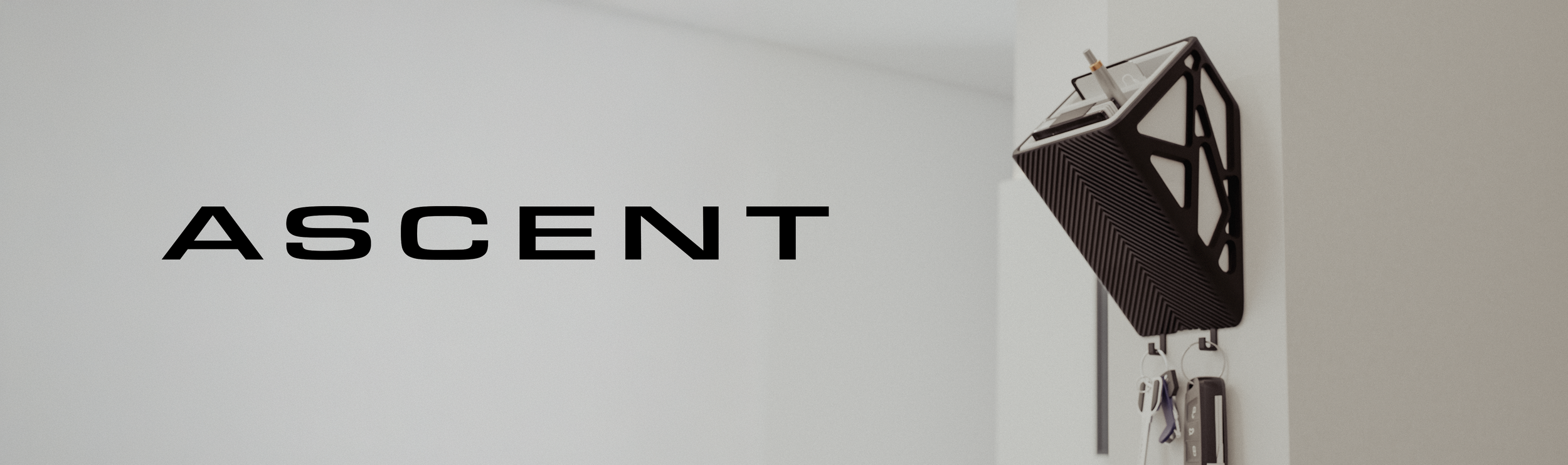 The wall displays the word 'ASCENT' in black capital letters with a black wall-mounted shelf holding a drink, sunglasses, and a keychain.