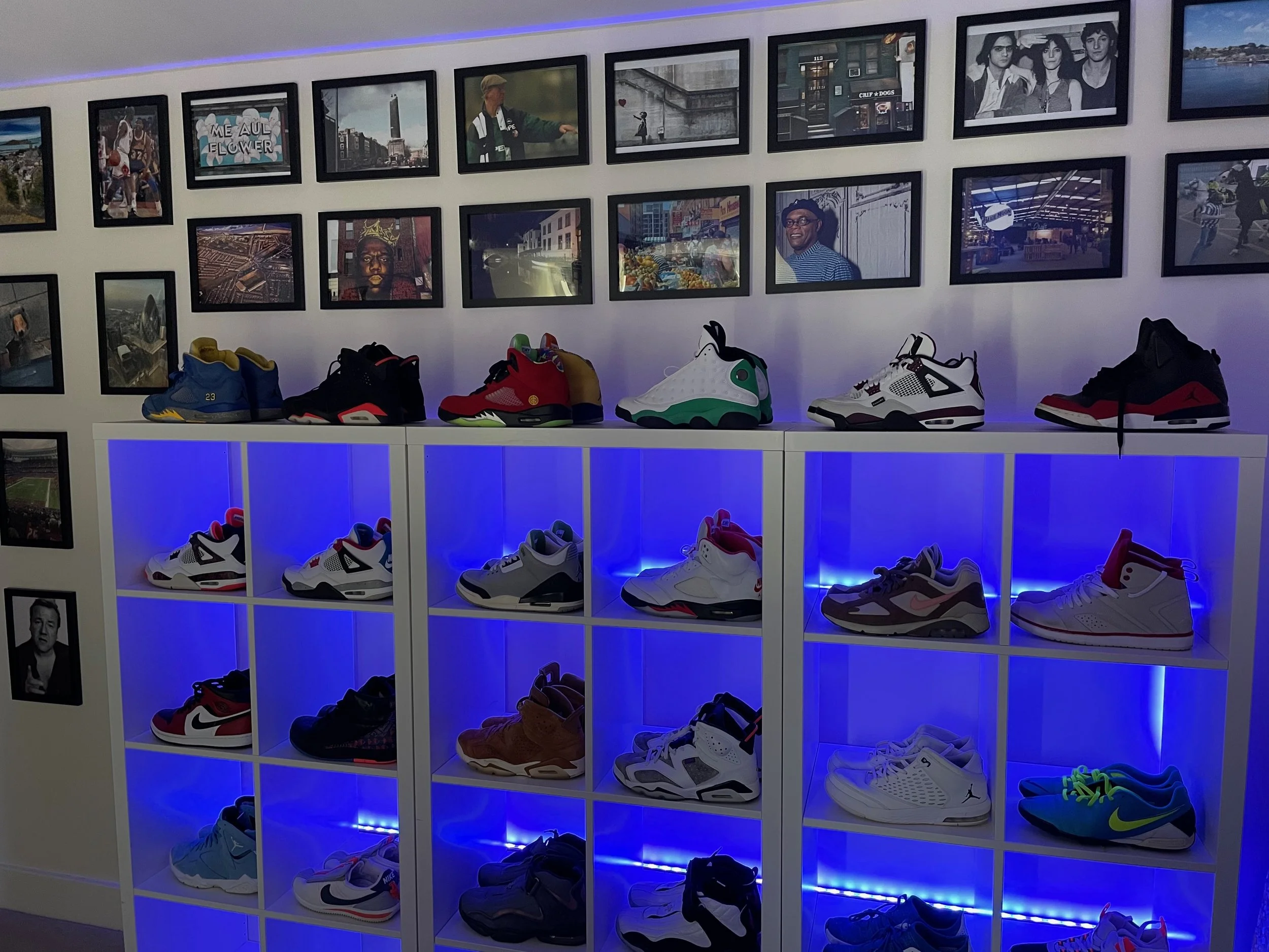 Display of various sneakers on white shelves with blue lighting, above a wall decorated with framed photos.