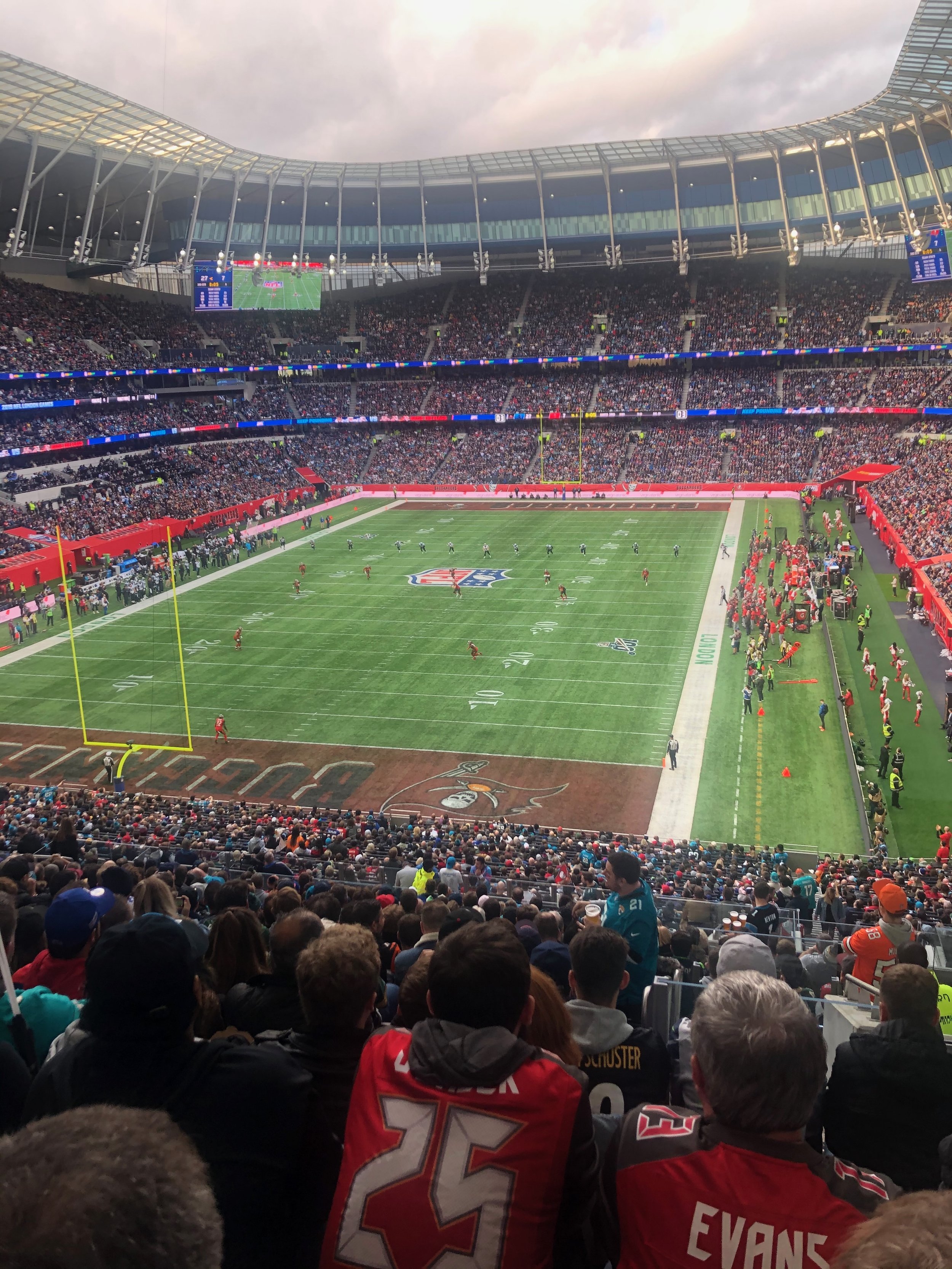 A football stadium filled with spectators, with players on the field warming up or playing. The field is marked with yard lines and the team logo in the center. The crowd covers multiple levels of the stadium, and the sky is cloudy.