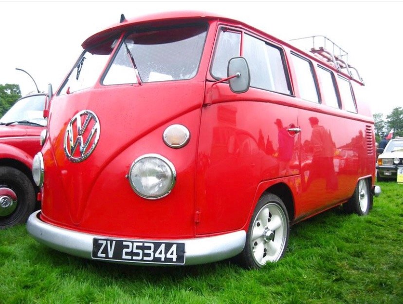 Red vintage Volkswagen Type 2 van with a split windshield parked on grass at a car show.