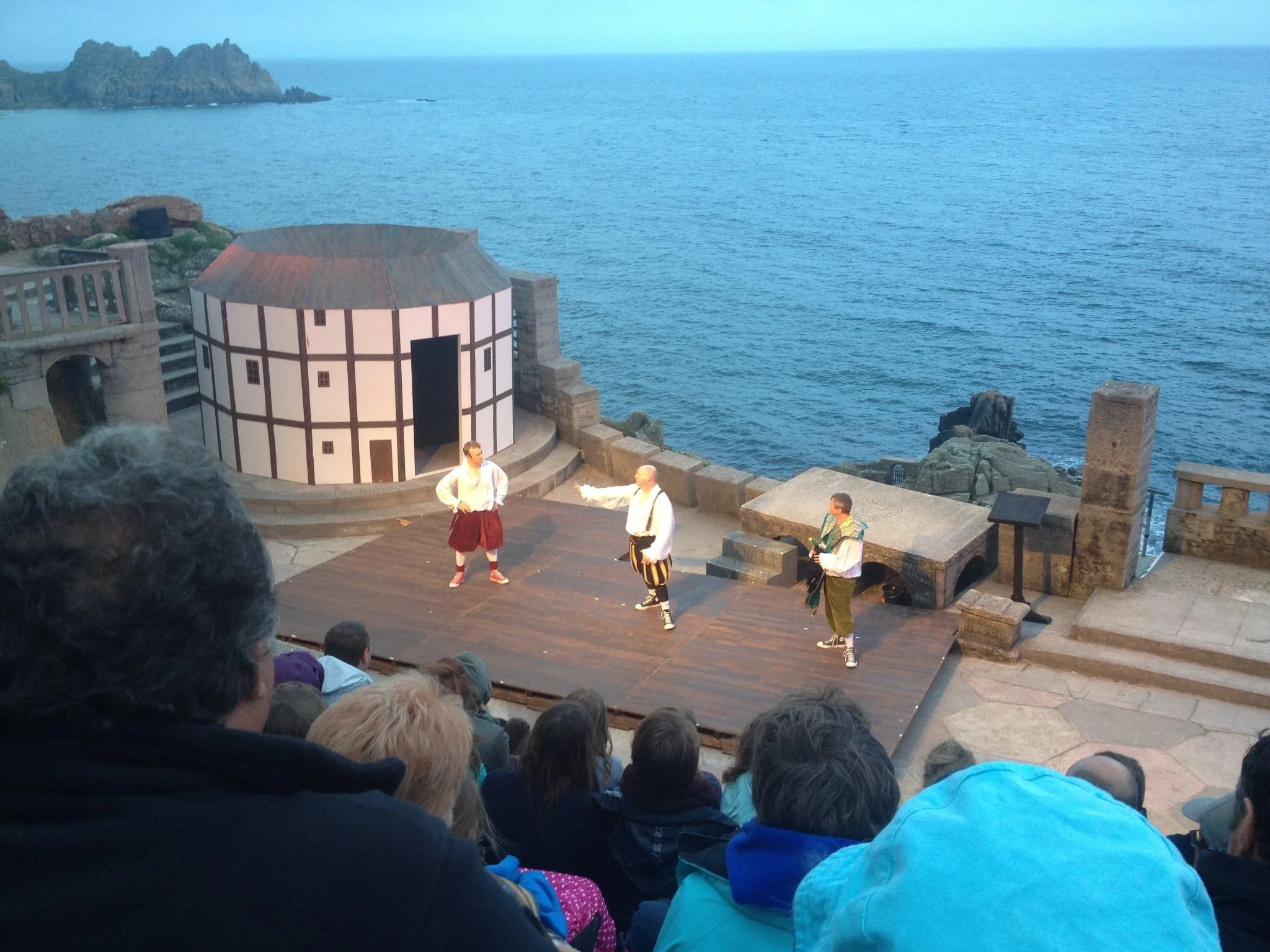 Performers in period costumes acting on an outdoor stage near the ocean, with an audience watching.
