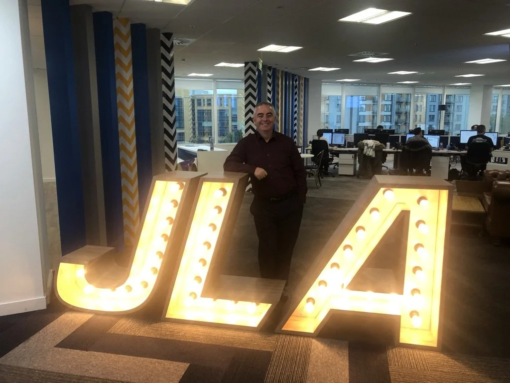 A man standing between illuminated large letter signs 'J' and 'A' in a modern office with workstations and large windows in the background.