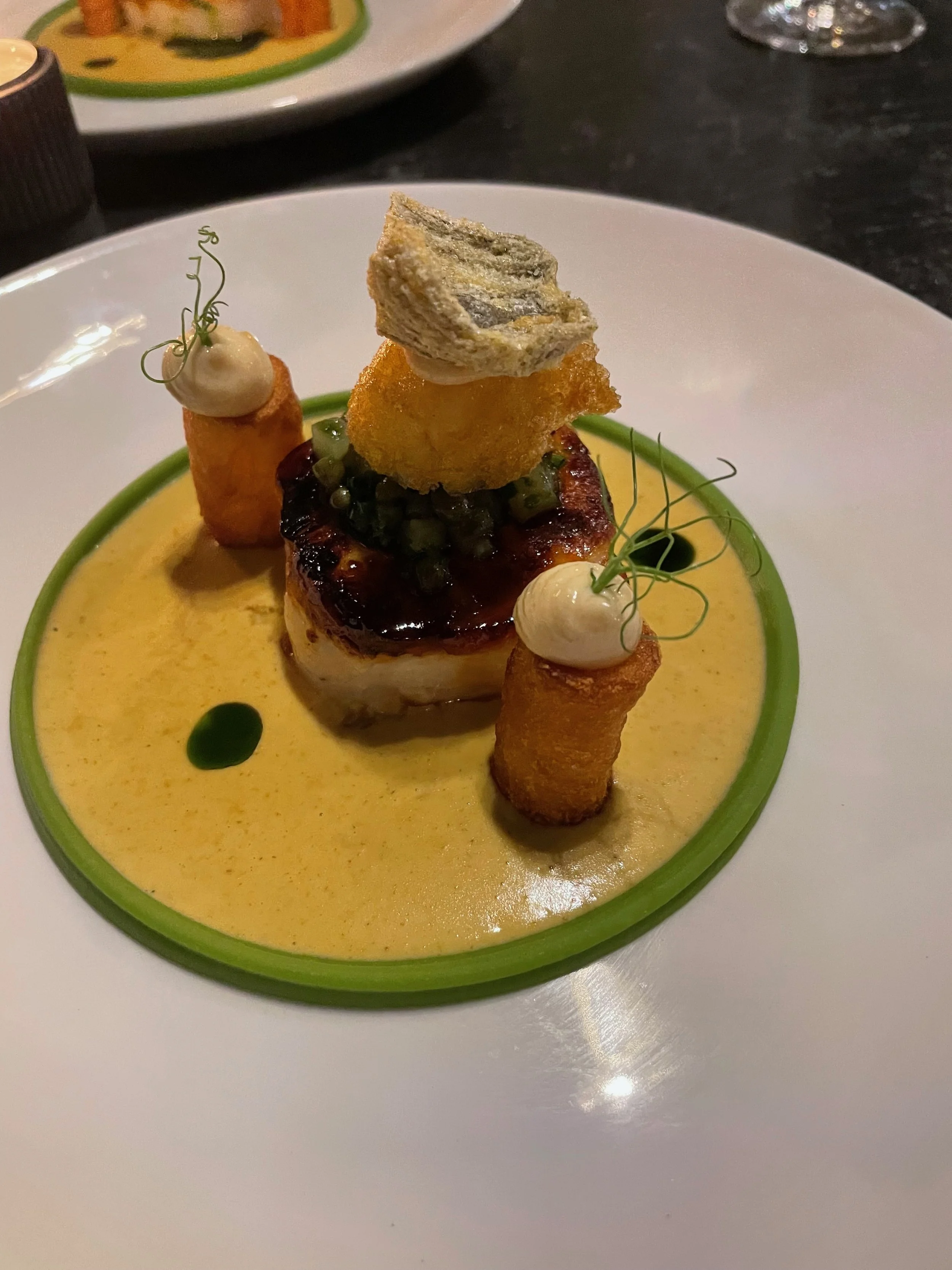 A gourmet plated dish with a central piece of seared meat topped with sauce and topped with fried vegetables, two rolled fried items with dollops of sauce, garnished with microgreens, presented on a white plate with a decorative green sauce ring.