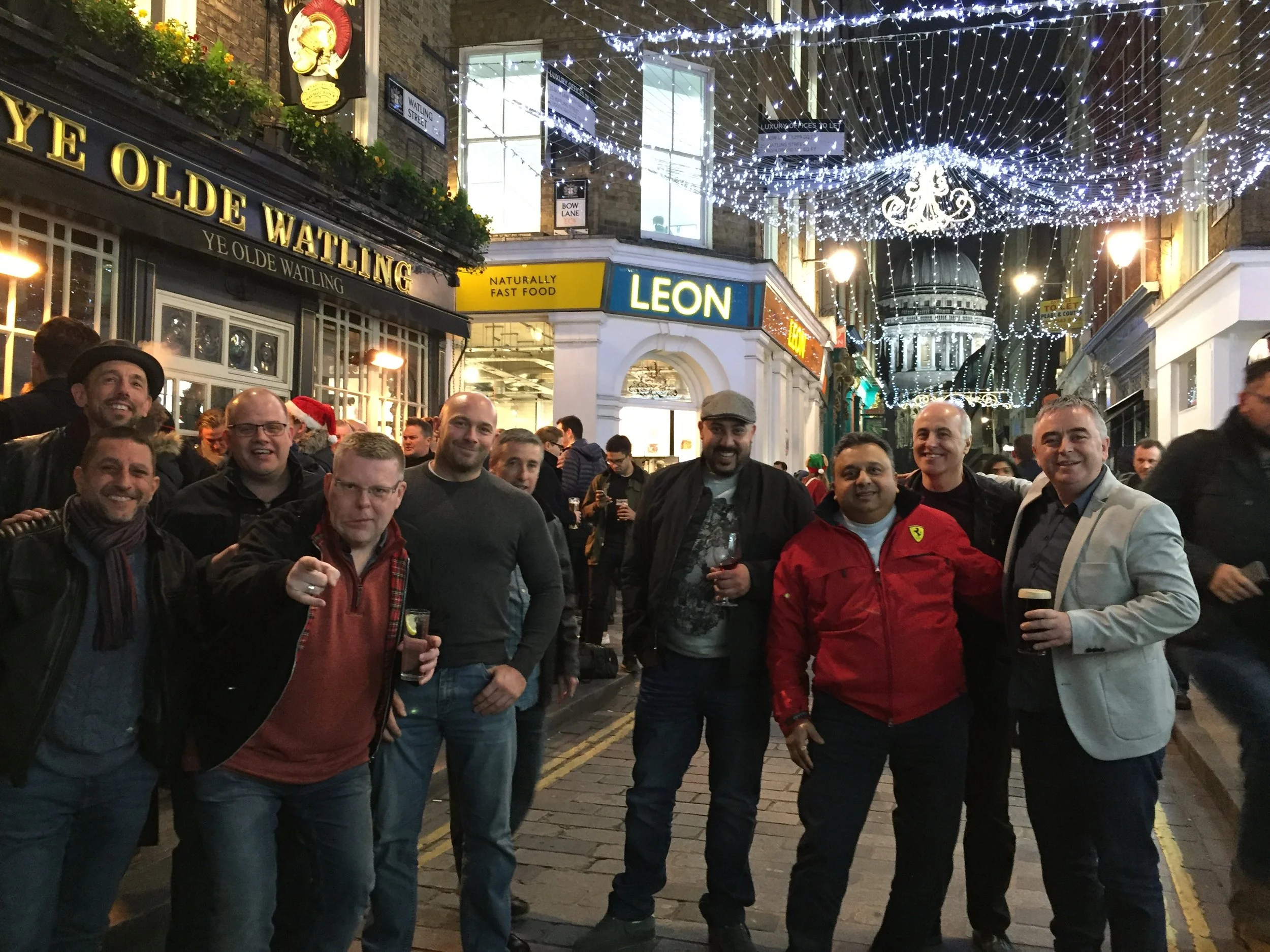 A group of nine men standing outdoors on a cobblestone street at night, smiling and holding drinks, with festive Christmas lights and decorations overhead, and storefronts nearby.