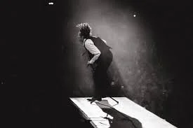 A person with long hair on a stage, bent over with their hands on their knees, standing next to a chair, in a dark, smoky setting.