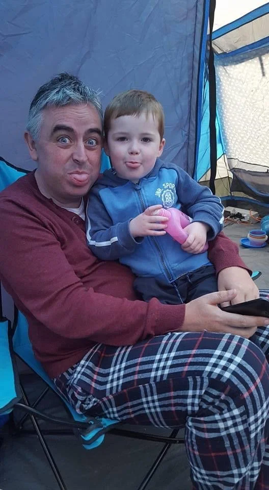 Adult man and young boy sitting together in a tent, making funny faces, with the man holding a remote control or phone.