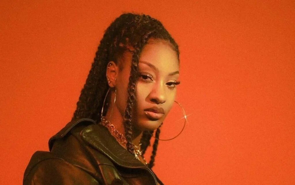 A young woman with long, styled braids, wearing hoop earrings and a leather jacket, standing against an orange background.