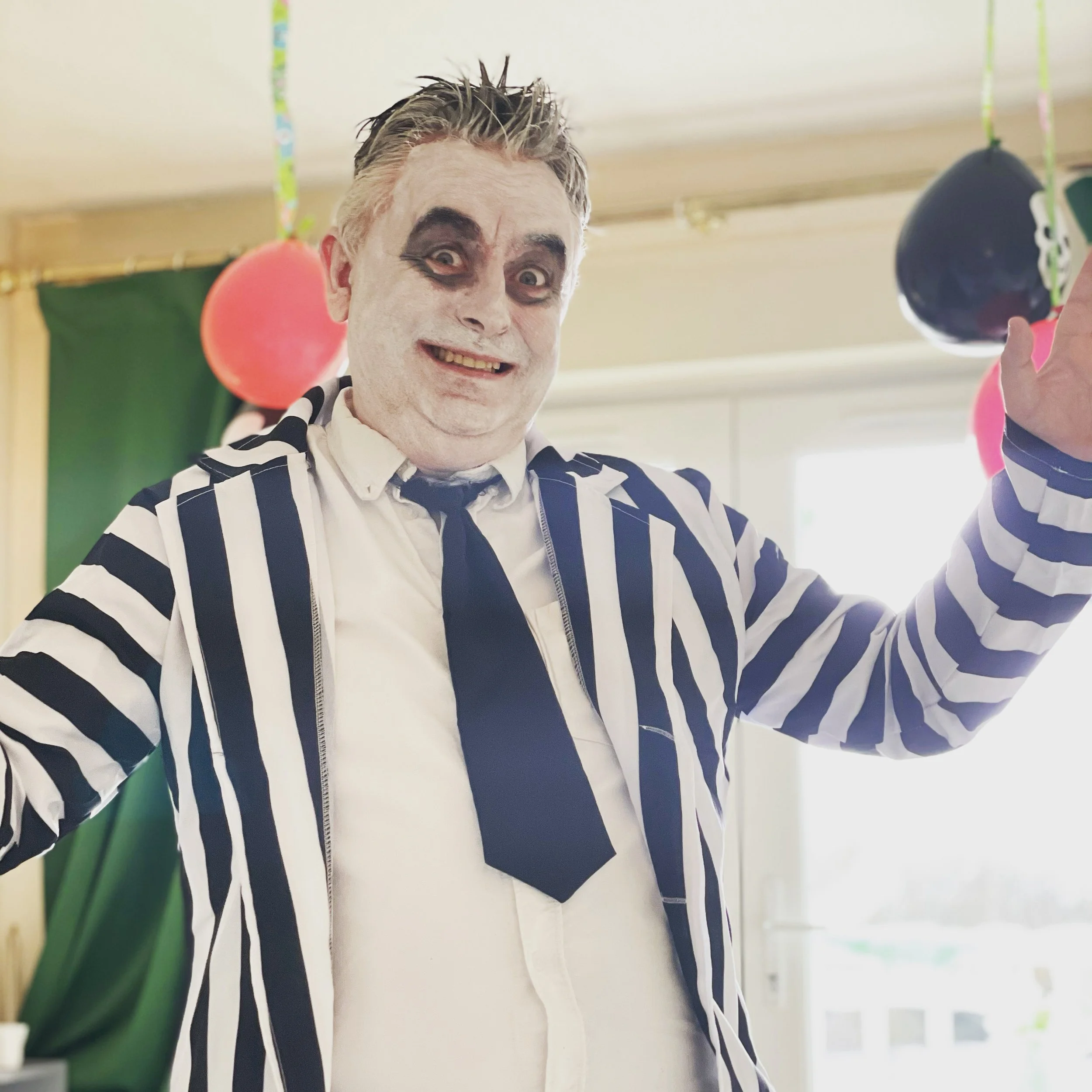 A person dressed as a clown or mime, with white face paint, black makeup around the eyes, and a big smile, wearing a black and white striped jacket and a black tie, celebrating at a party with balloons in the background.