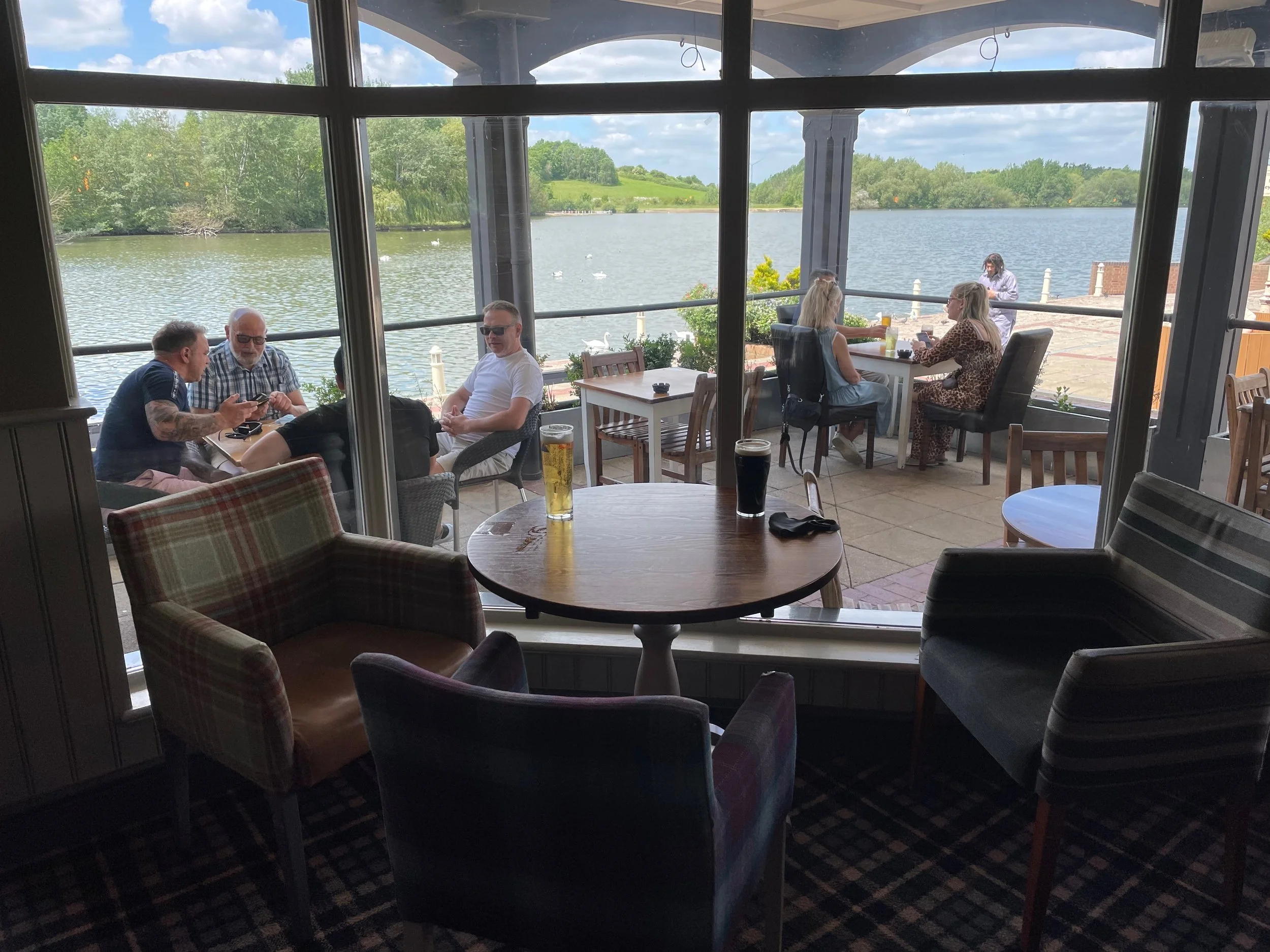 People sitting outside by a lake, enjoying drinks and conversation on a patio with large windows offering a view of the water and trees.
