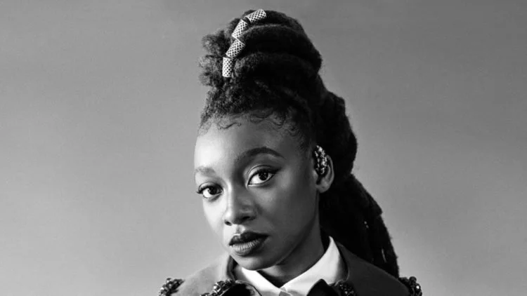 Black woman with styled dreadlocks, wearing a bow hair accessory and a collared shirt, looking directly at the camera in black and white.