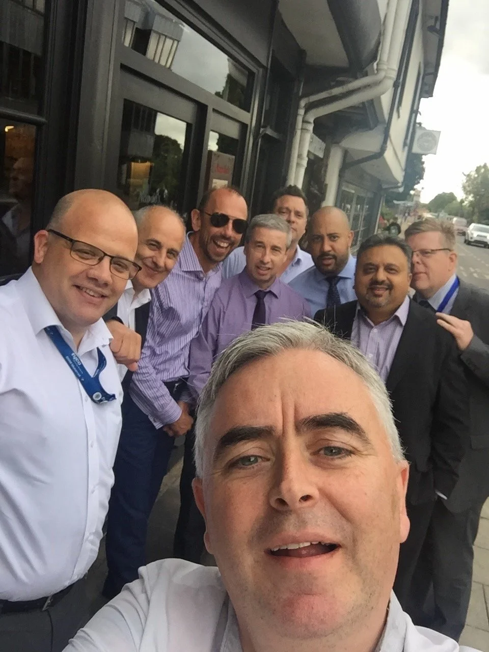 A group of nine men smiling for a selfie outside a building on a city street.