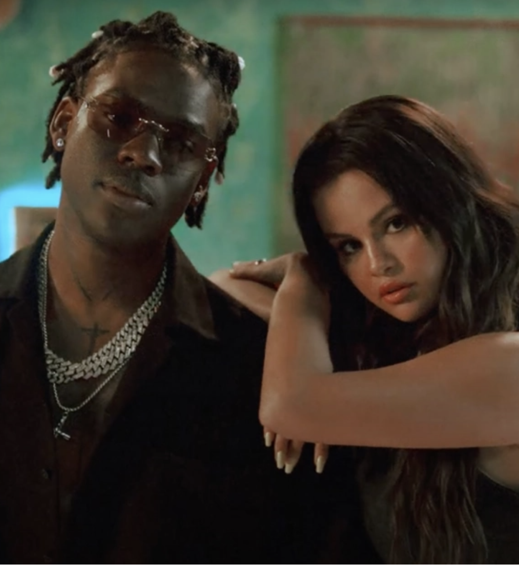Two young adults, a man with sunglasses, jewelry, and dreadlocks, and a woman with long dark hair, pose closely together in a dimly lit environment.