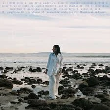Person standing on a rocky beach near the ocean, wearing white clothing, looking out at the water.