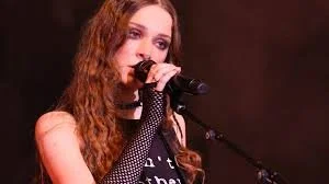 Young woman with long, wavy hair singing into a microphone on stage.