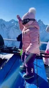 Child in pink winter jacket, beanie, and sunglasses skiing in snowy mountains.