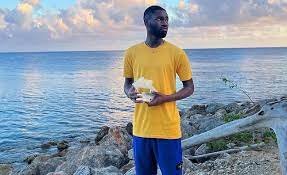 A man in a yellow shirt and blue shorts standing near the ocean, holding a pizza box, with a rocky shoreline and boat in the background.