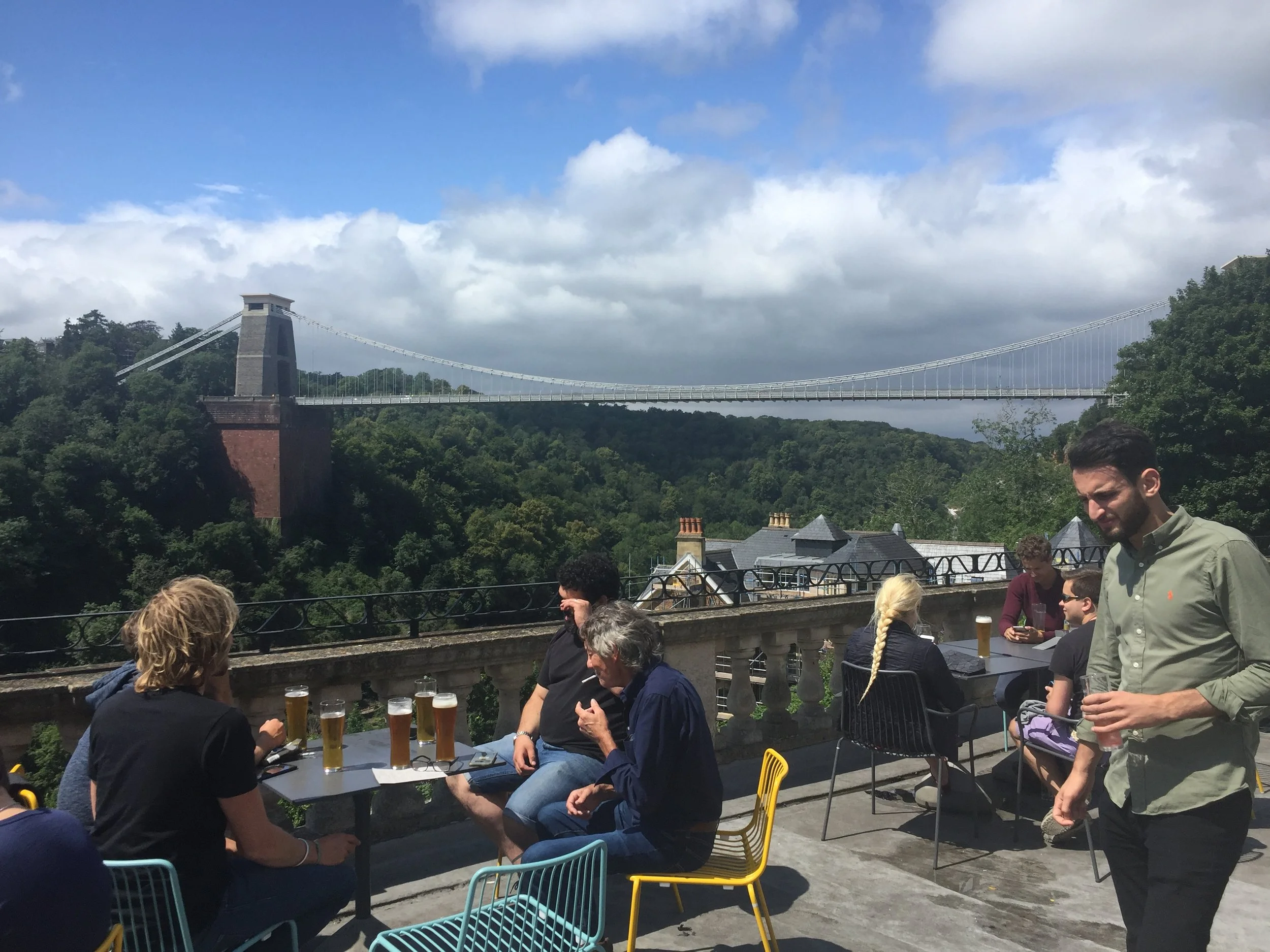 People sitting at an outdoor rooftop bar with glasses of beer, overlooking a bridge spanning a green valley, under a partly cloudy sky.