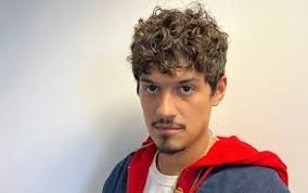A young man with curly hair and a light beard, wearing a red jacket over a white shirt, looking at the camera against a plain background.