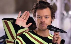 A man with dark, curly hair holding a microphone in his right hand and waving with his left hand.