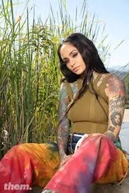A woman with tattoos sitting outdoors among tall green plants, wearing a sleeveless tan top and colorful pants.