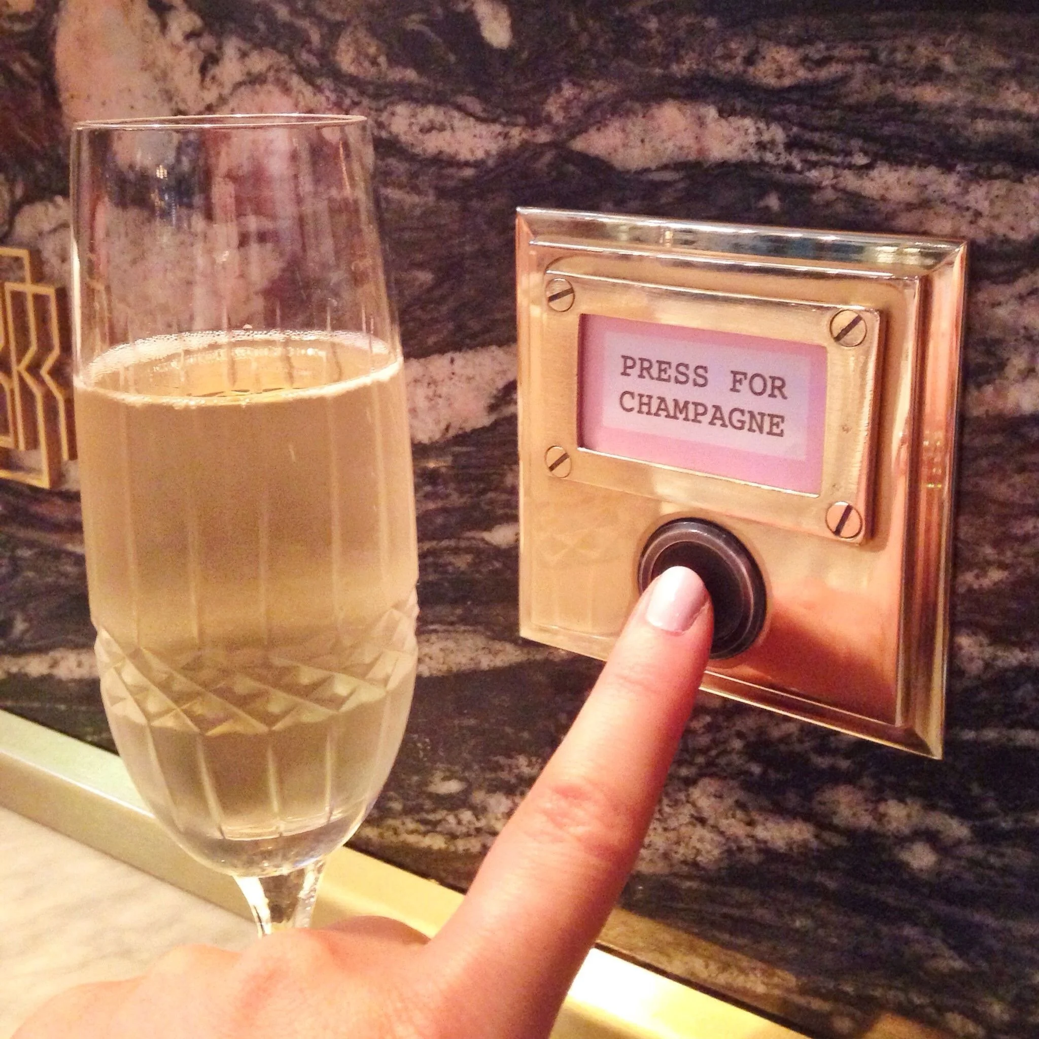 A hand pressing a button labeled "Press for Champagne" next to a glass filled with champagne on a marble counter.