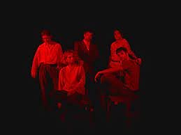 Five people posing against a black background, illuminated with red lighting, some sitting and others standing.