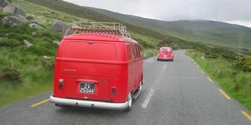 Red vintage van and pink Volkswagen Beetle driving on a scenic mountain road with green hills and rocks
